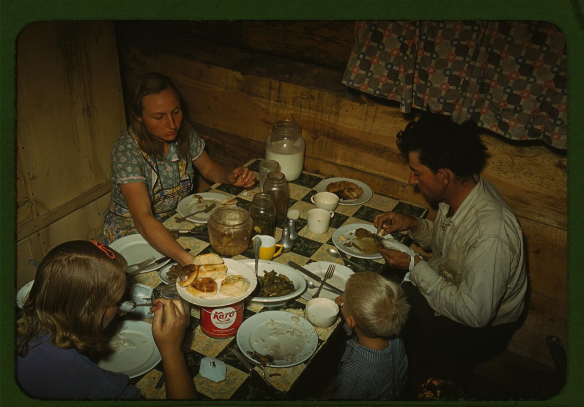 A family of four eating dinner at a table set with plates, jars and a lamp inside a log-walled dugout home, in color.