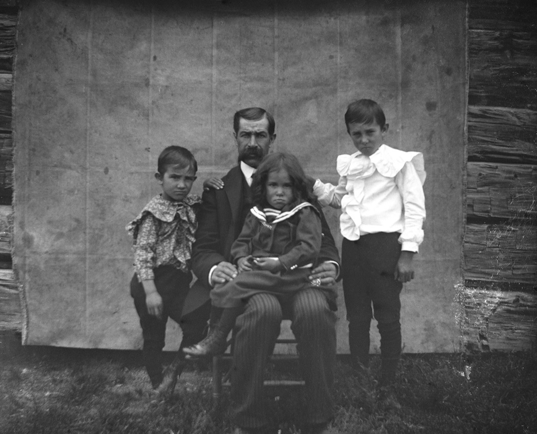 A father seated with his young daughter on his lap and two sons standing beside him, in front of a cloth backdrop hung on a log wall, about 1900.